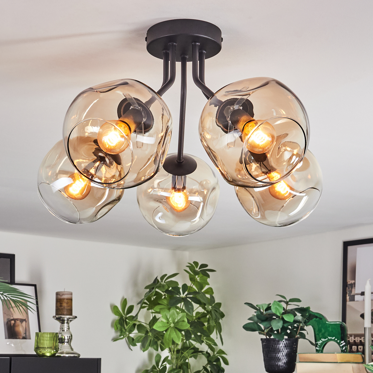 Eine moderne Deckenleuchte mit fünf bernsteinfarbenen Glaskugeln, in denen sich jeweils eine warme Glühbirne befindet. Schwarze Metallbefestigungen. Pflanzen und Dekoration im Hintergrund.