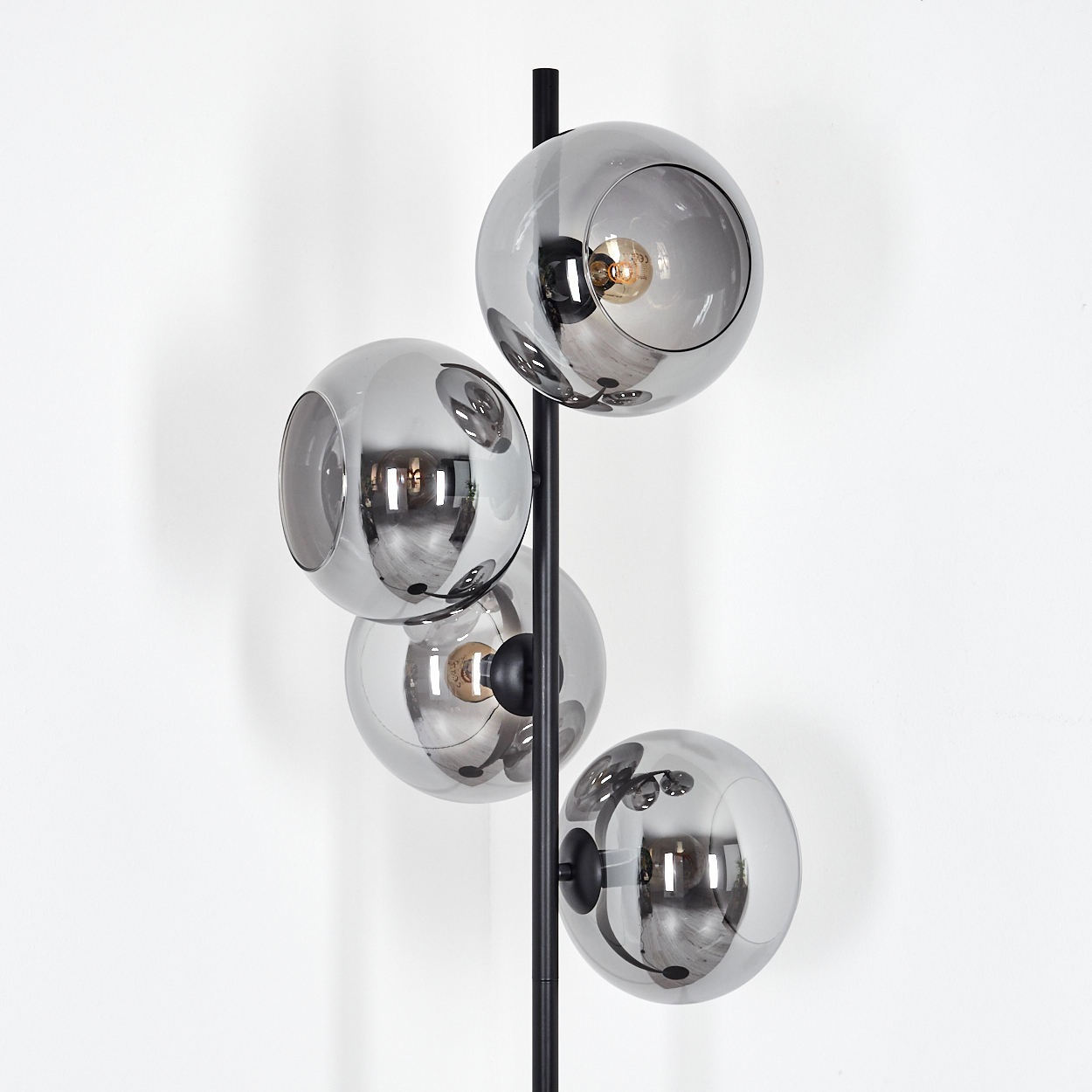 Eine moderne Stehlampe mit vier grauen Glaskugelschirmen. Die Lampe hat ein schwarzes Metallgestell vor einer weißen Wand.