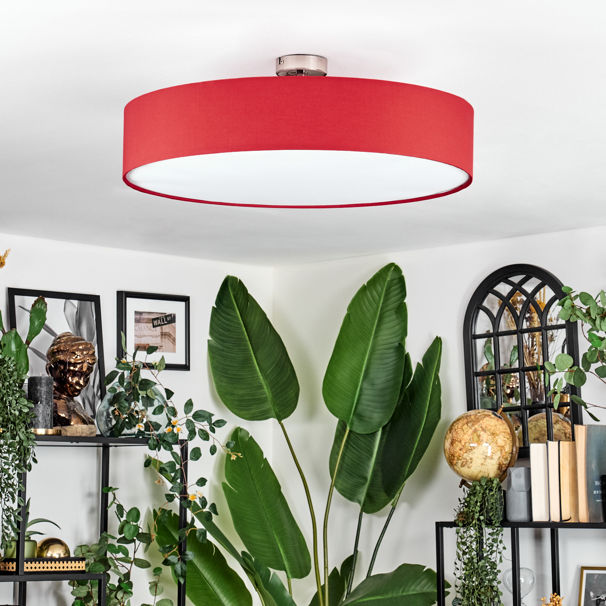 Eine rote Deckenlampe hängt über einem Raum mit Pflanzen und Regalen. Ein Globus und Bücher sind sichtbar. Große grüne Blätter sind im Vordergrund.