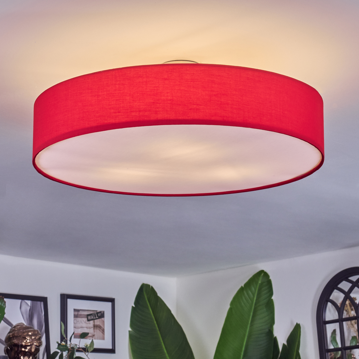 Eine große rote Deckenlampe ist der Mittelpunkt. Im Vordergrund sind grüne Pflanzen und gerahmte Bilder zu sehen.