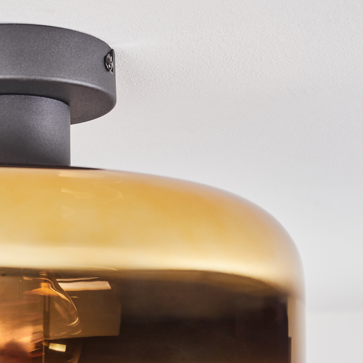 Nahaufnahme einer Deckenlampe. Sie hat einen abgerundeten, bernsteinfarbenen Glasschirm und einen dunklen Metallsockel. Der Hintergrund ist eine weiße Decke.