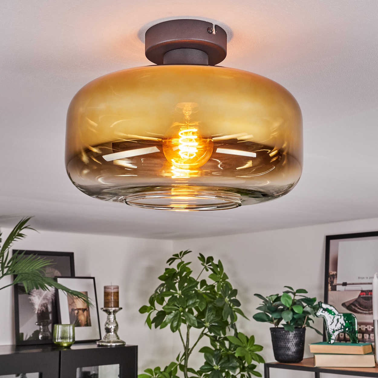 Eine moderne Deckenlampe mit braunem Sockel und bernsteinfarbenem Glasschirm. Die Glühbirne leuchtet und beleuchtet den Raum, darunter Pflanzen und Dekoration.