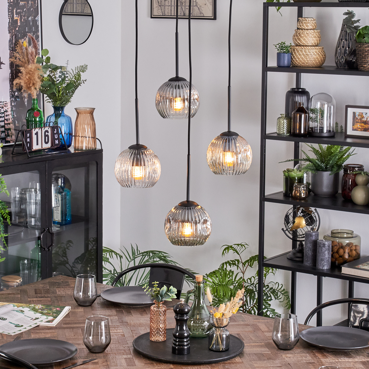 Esszimmer mit Holztisch, Hängelampen, schwarzen Regalen mit Deko und Pflanzen. Tisch gedeckt mit Tellern und Gläsern.