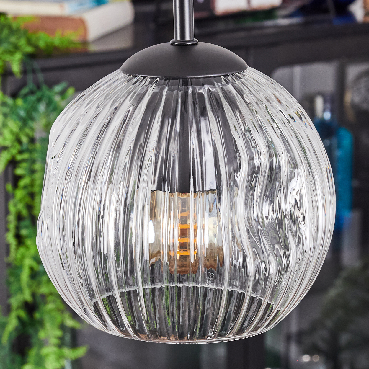 Eine klare, gerippte Glas-Pendelleuchte hängt von der Decke. Die Glühbirne ist sichtbar. Im Hintergrund sind grüne Pflanzen zu sehen.