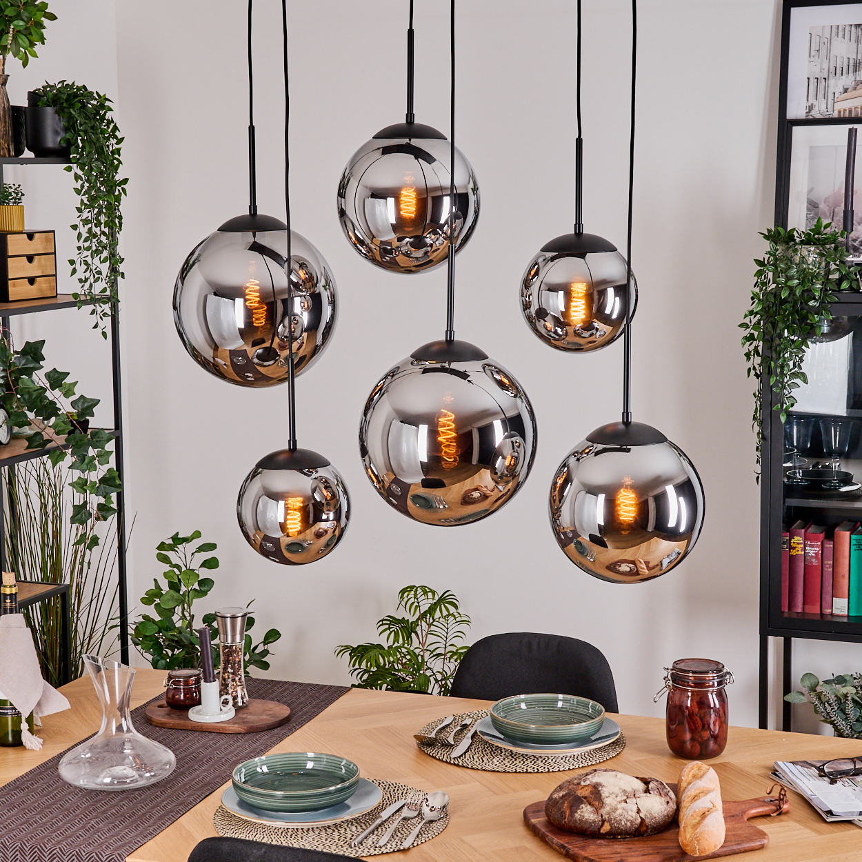 Esszimmer mit sechs runden Chrom-Hängelampen. Tisch gedeckt mit Tellern, Brot und einem Glas mit Essen. Pflanzen umgeben die Szene.