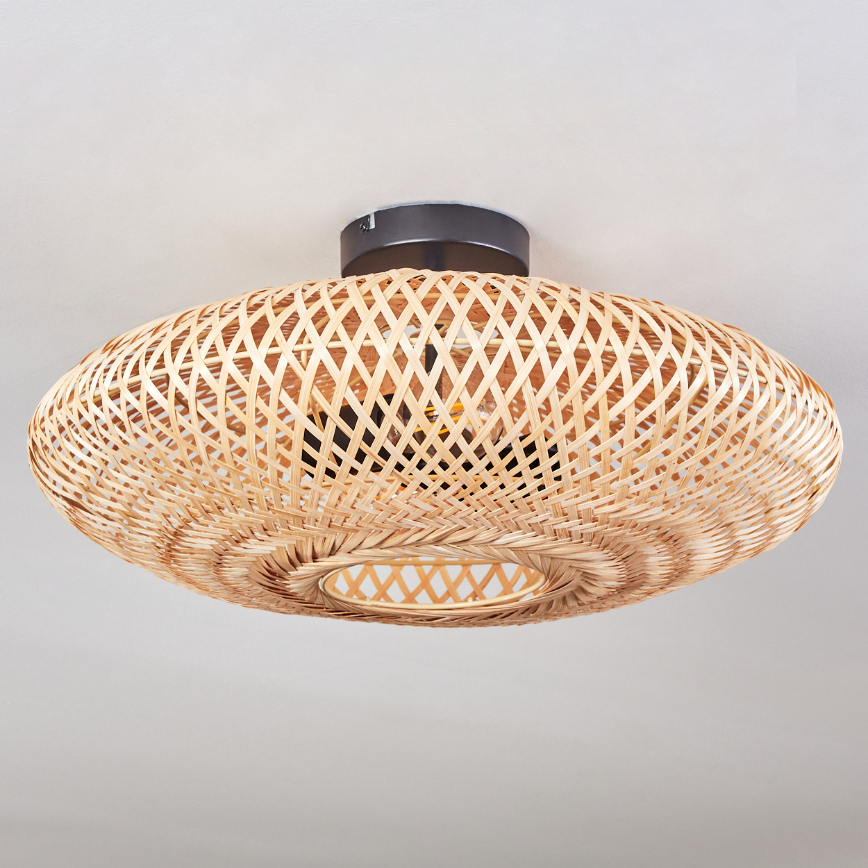Eine Deckenlampe aus geflochtenem Rattan. Die Lampe ist oval mit einem dunklen Metallsockel. Die Glühbirne im Inneren ist sichtbar und beleuchtet das Webmuster.