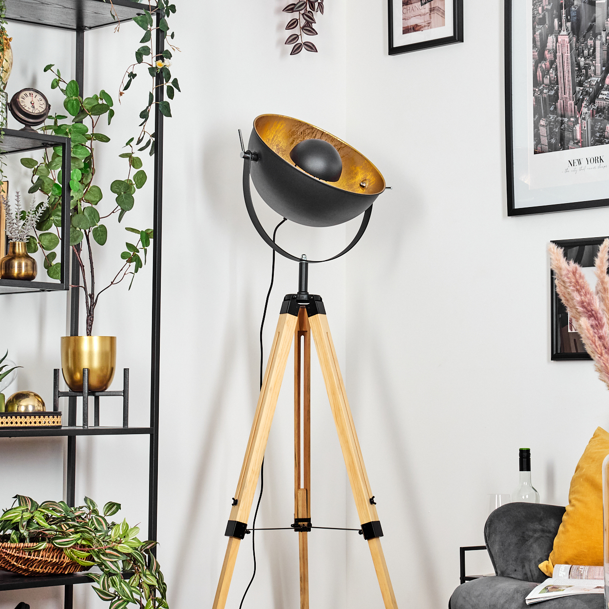 Eine schwarz-goldene Stehlampe mit Holzstativ. Pflanzen und Regale im Hintergrund, zusammen mit einem gerahmten Bild.