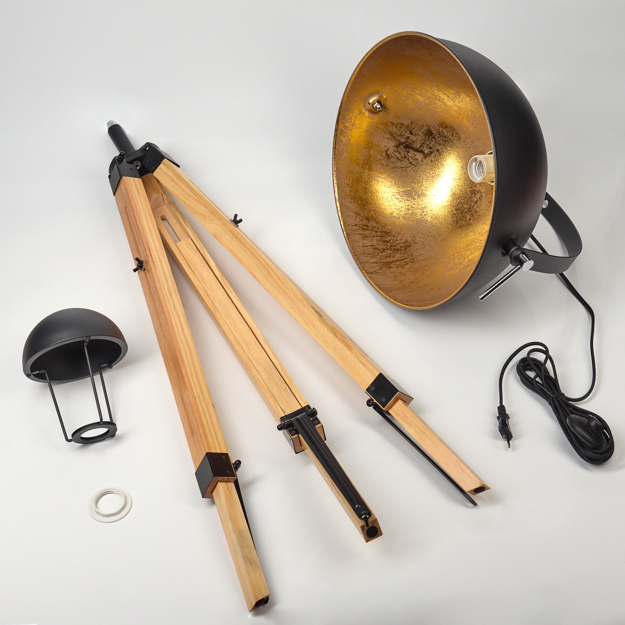 Eine zerlegte schwarz-goldene Lampe mit hölzernen Stativbeinen, einem Lampenschirm und einem Netzkabel, alles auf einer weißen Oberfläche.