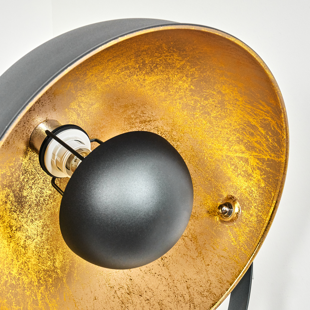 Nahaufnahme einer Lampe. Schwarzer Schirm mit goldenem Innenraum und sichtbarer Glühbirne. Der Hintergrund ist weiß.