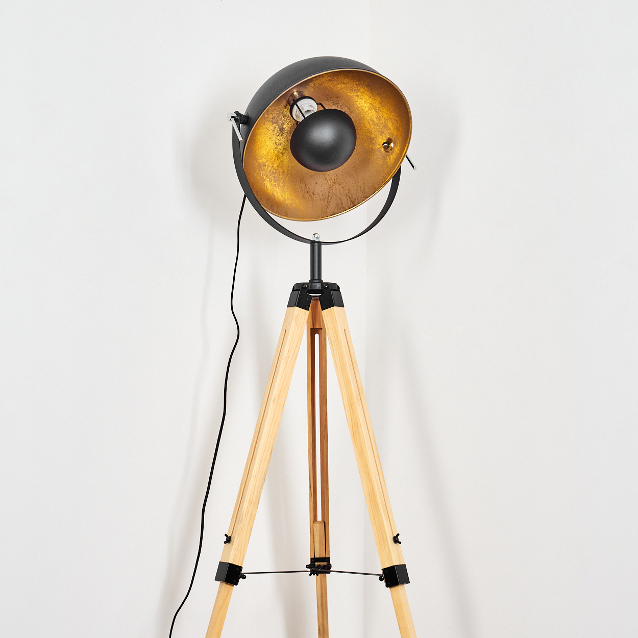 Eine Stehlampe mit schwarzem und goldenem Lampenschirm, Holz-Dreibein-Beinen und einem schwarzen Draht vor weißer Wand.