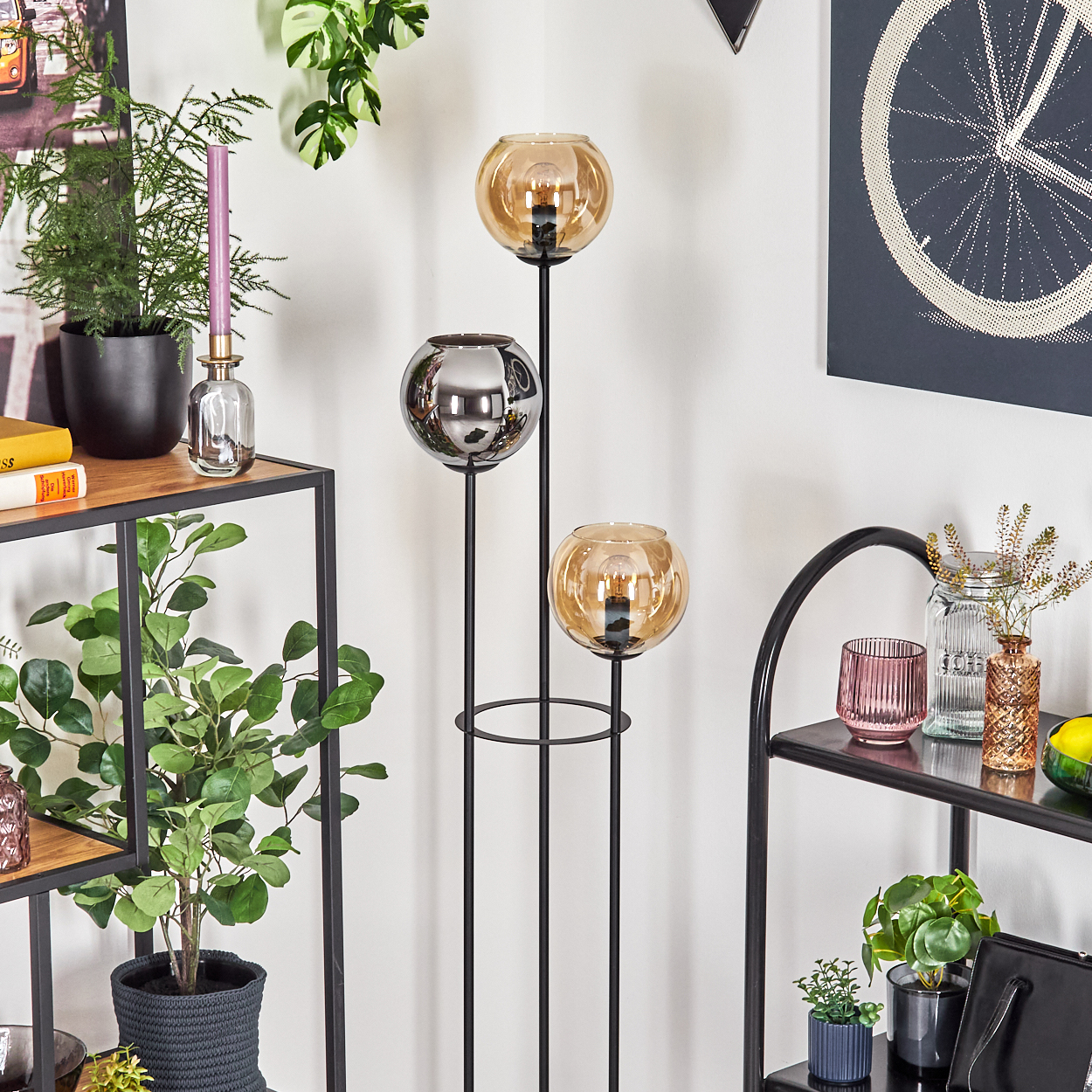 Ein modernes Zimmer mit einem schwarzen Metallregal, Pflanzen und einer Dreifach-Glühbirnenlampe mit Glaskugeln. An der Wand befindet sich ein Fahrradrad-Kunstwerk.