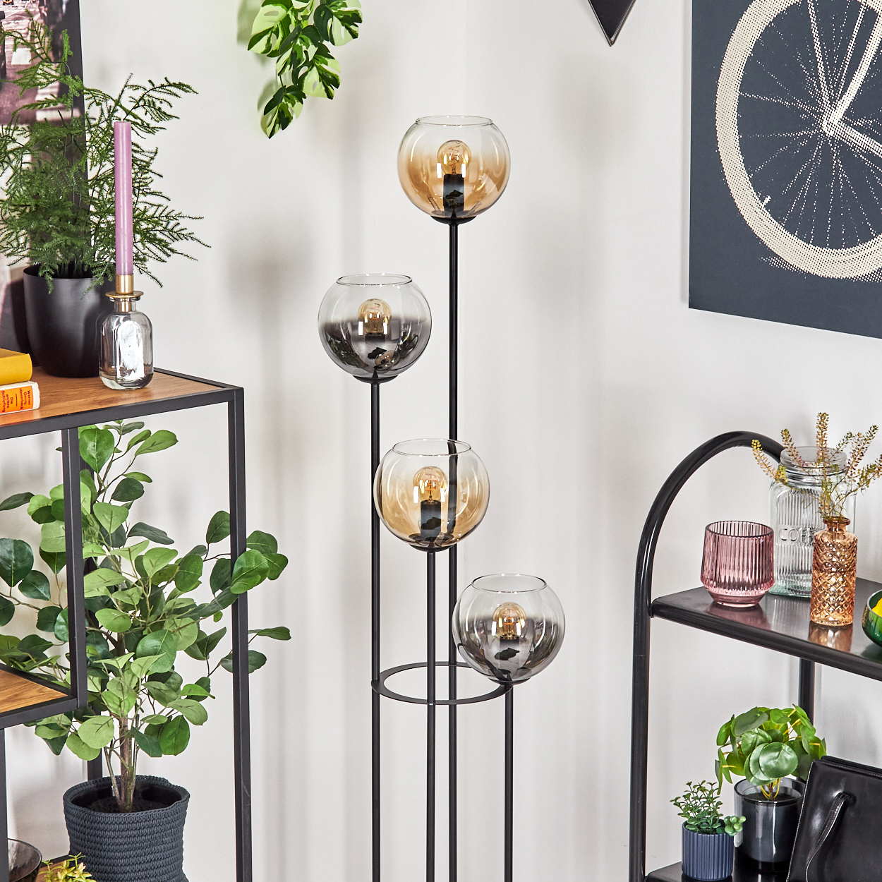 Eine moderne Stehlampe mit vier Glaskugeln, jede mit einer Glühbirne. Es ist in einem Raum mit Pflanzen und Regalen.