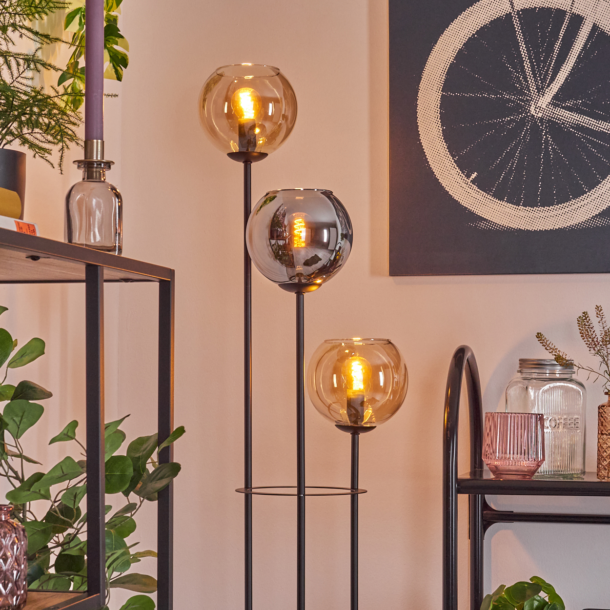 Eine dreiflammige Lampe mit bernsteinfarbenen Glaskugeln. Im Hintergrund ein Regal mit Pflanzen, Fahrradrad-Kunst und anderen dekorativen Gegenständen.