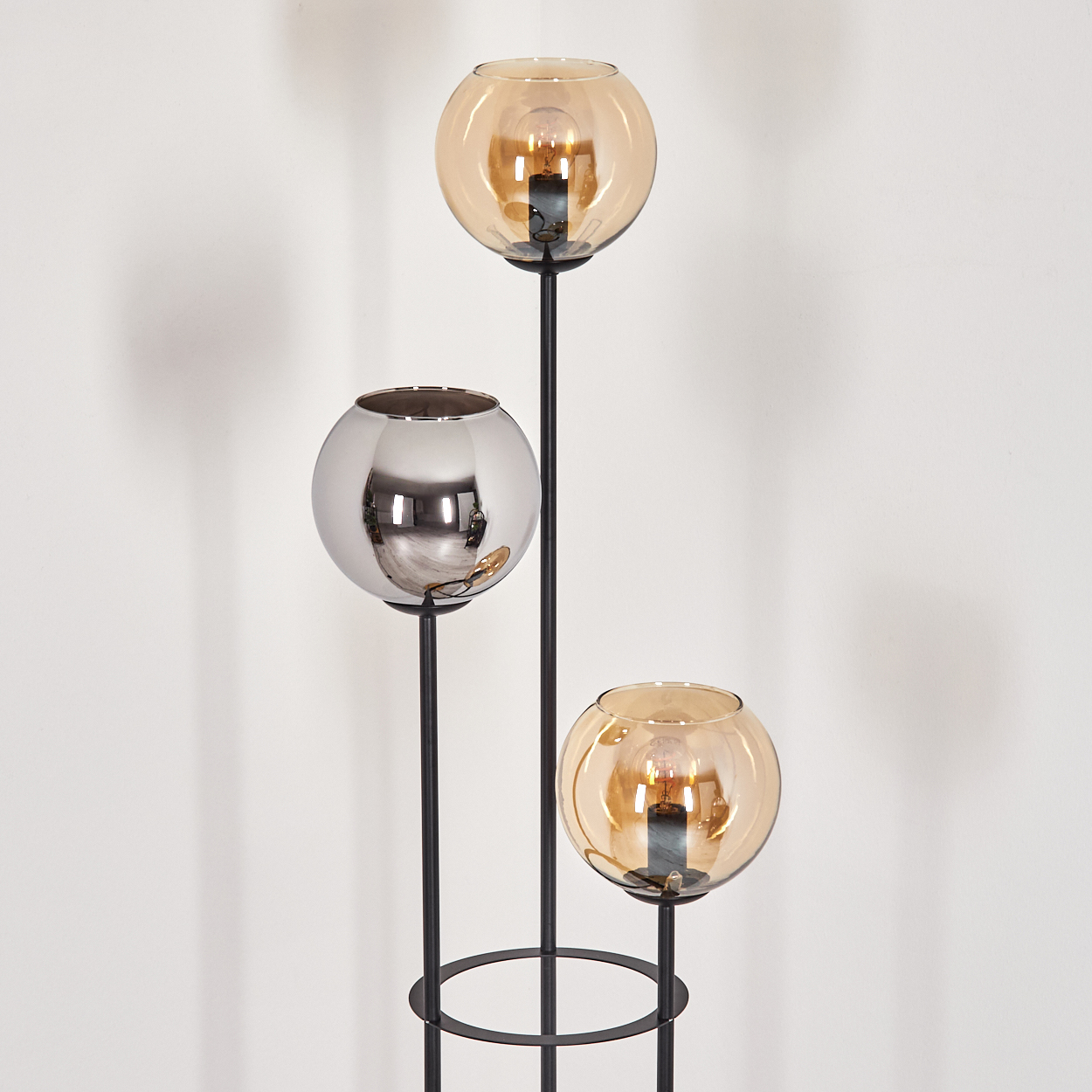 Eine moderne Lampe mit drei kugelförmigen Schirmen. Zwei bernsteinfarbene Schirme mit leuchtenden Glühbirnen, ein Chromschirm. Schwarzer Rahmen, weißer Hintergrund.