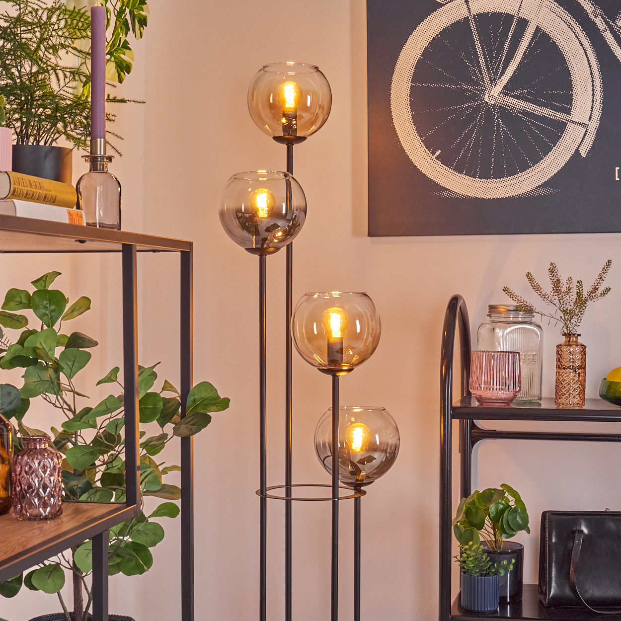 Ein modernes Wohnzimmer mit einer Stehlampe mit vier Glaskugeln. Regale mit Pflanzen, Kerzen und Dekoration sowie ein Fahrradkunstwerk.