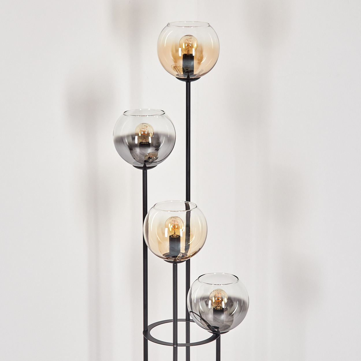 Eine moderne Stehlampe mit vier Glaskugelschirmen, jede mit einer Glühbirne. Die Lampe ist schwarz, der Hintergrund ist eine weiße Wand.