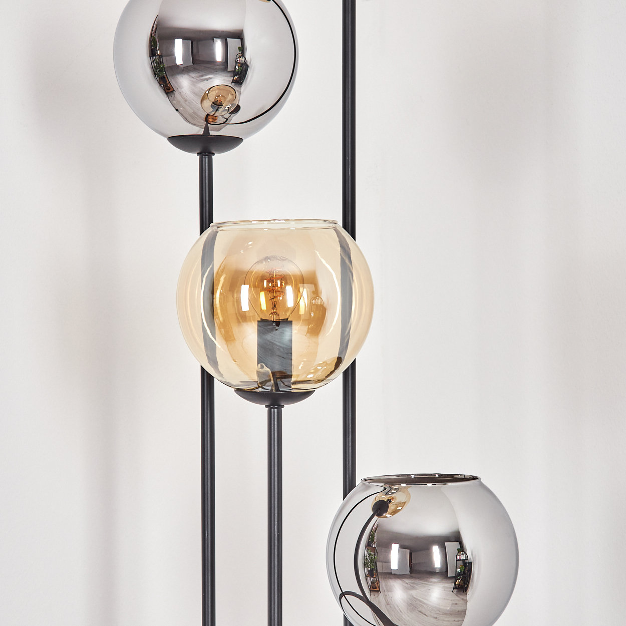 Moderne Lampe mit drei kugelförmigen Schirmen. Zwei sind verspiegelt, einer bernsteinfarben. In jeder Kugel ist eine Glühbirne zu sehen. Schwarzer Rahmen.