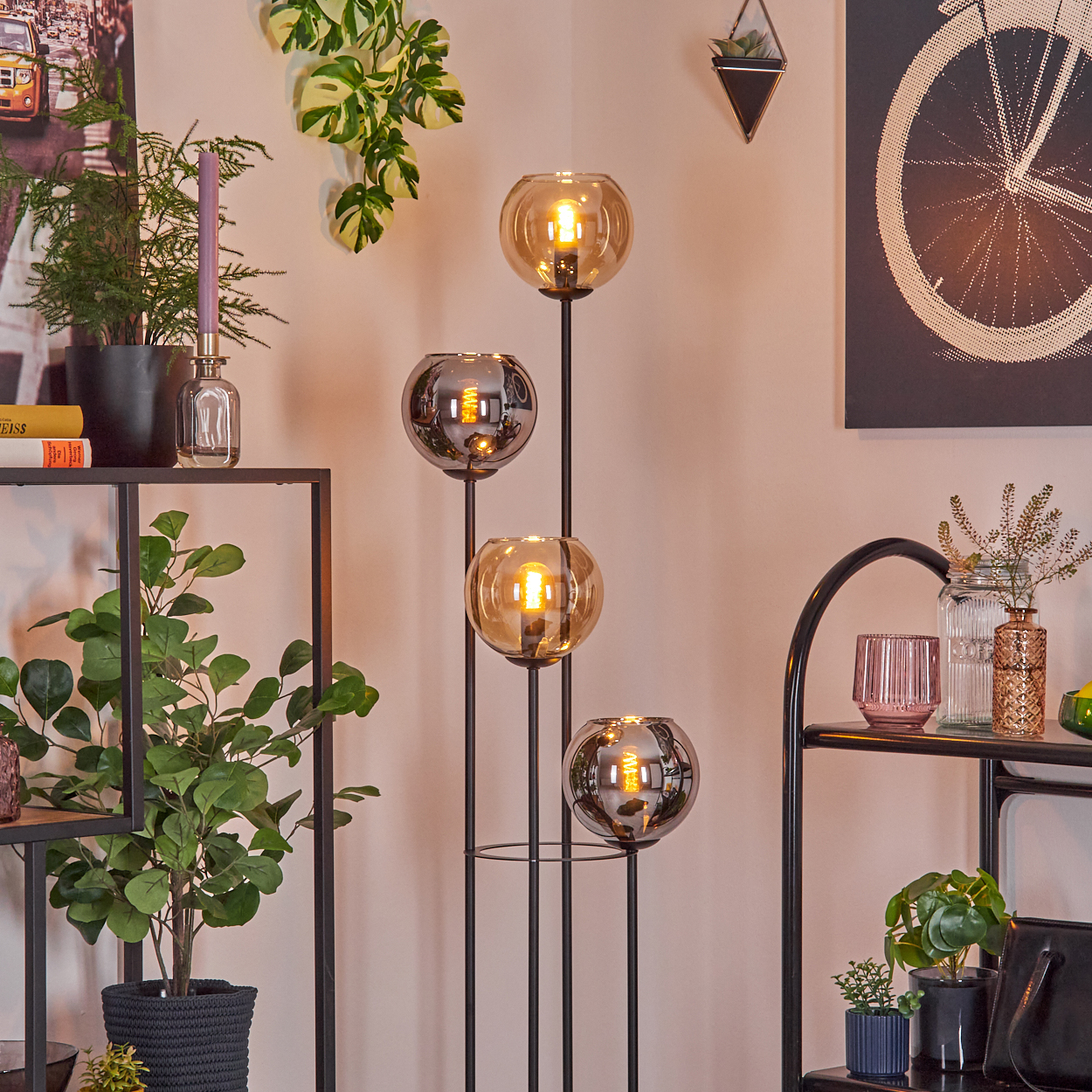 Eine moderne Stehlampe mit vier Glaskugeln. Regale mit Pflanzen und Dekor befinden sich im Vordergrund. Ein Fahrradrad-Kunstwerk ist an der Wand.