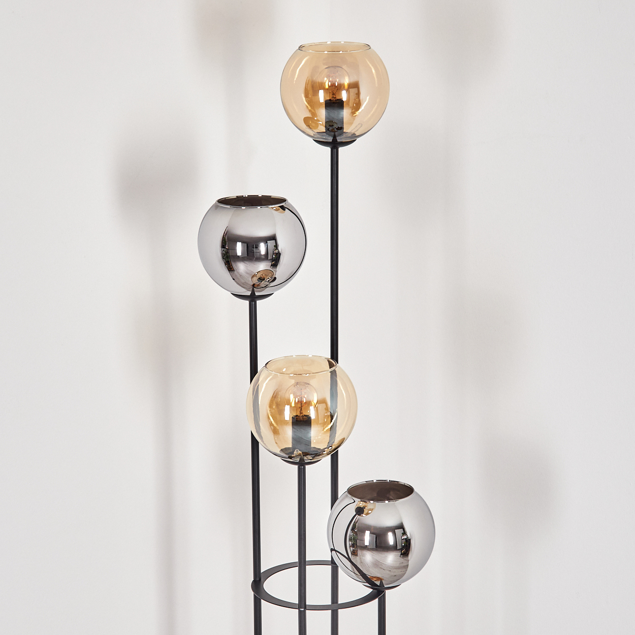 Eine moderne Lampe mit vier Glasschirmen in Bernstein und Silber, auf schwarzen Metallstielen, vor einer weißen Wand.