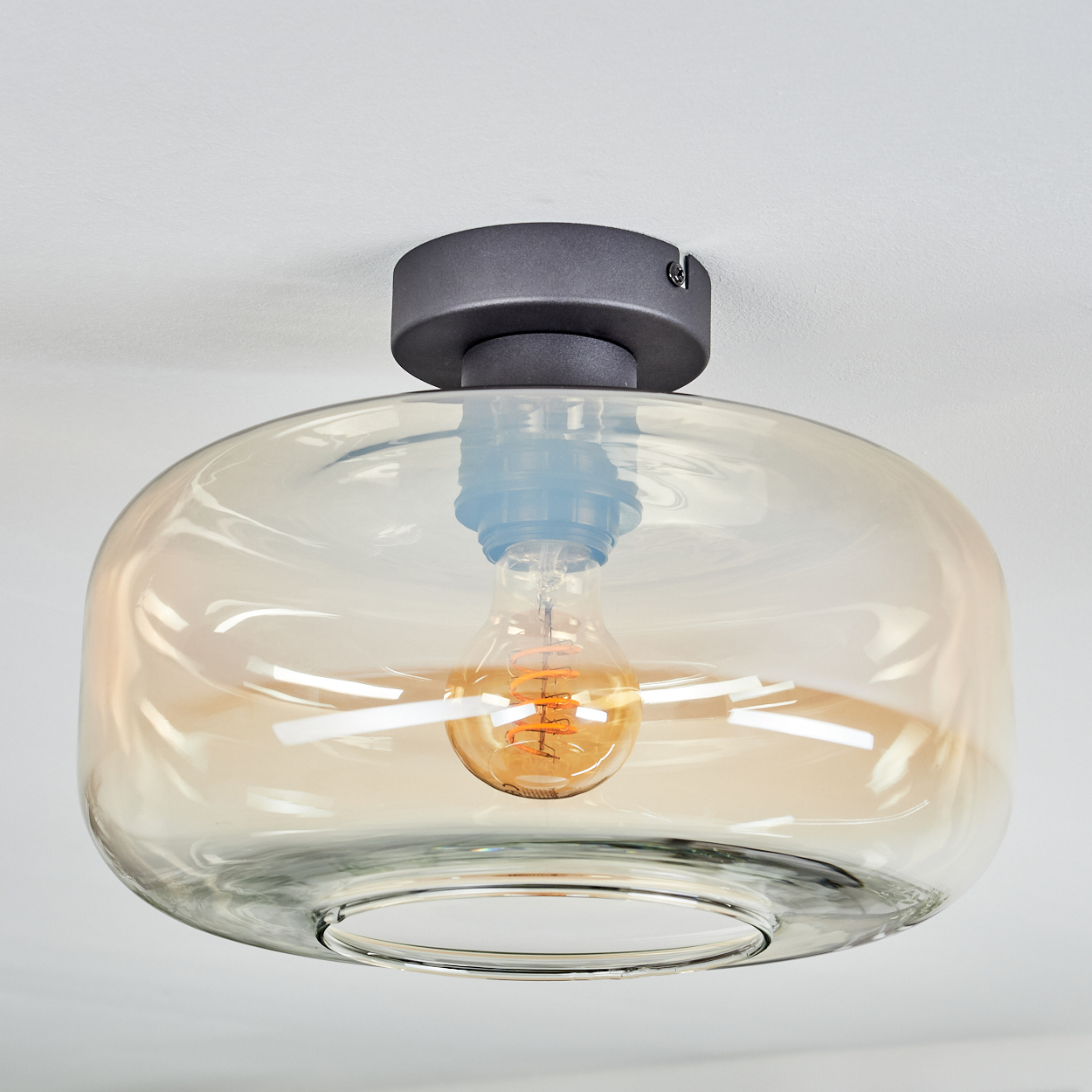 Eine moderne Deckenlampe mit klarem Glasschirm und sichtbarer orangefarbener Glühbirne. Die Leuchte ist an einer weißen Decke befestigt.