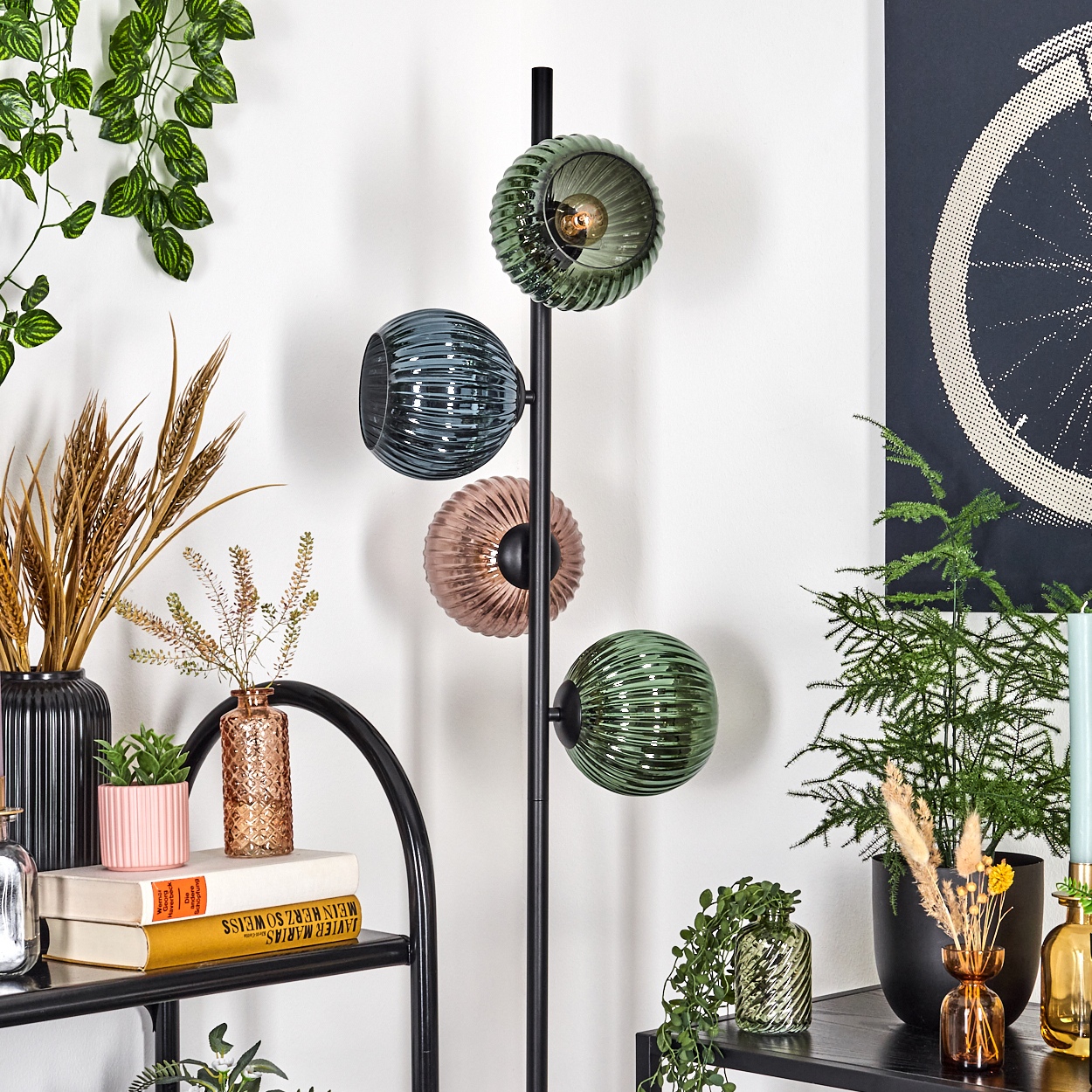 Eine moderne Stehlampe mit drei Glaskugeln. Verschiedene Pflanzen und Dekorationsartikel auf Regalen und dem Boden, vor einer weißen Wand.