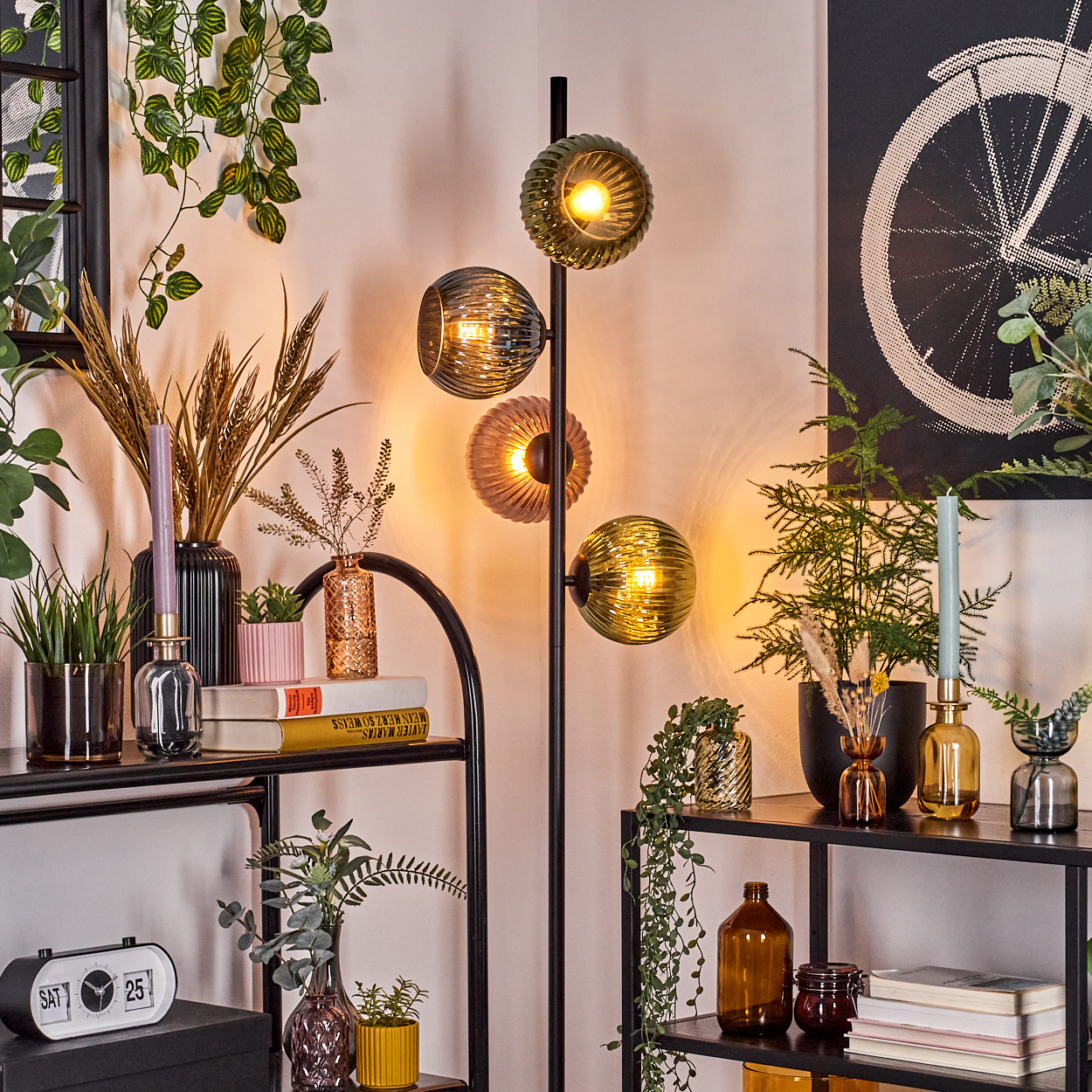Ein Raum mit einer Stehlampe und Regalen. Die Regale beherbergen Pflanzen, Bücher und dekorative Vasen. Im Hintergrund hängt ein Fahrradrad-Kunstwerk.