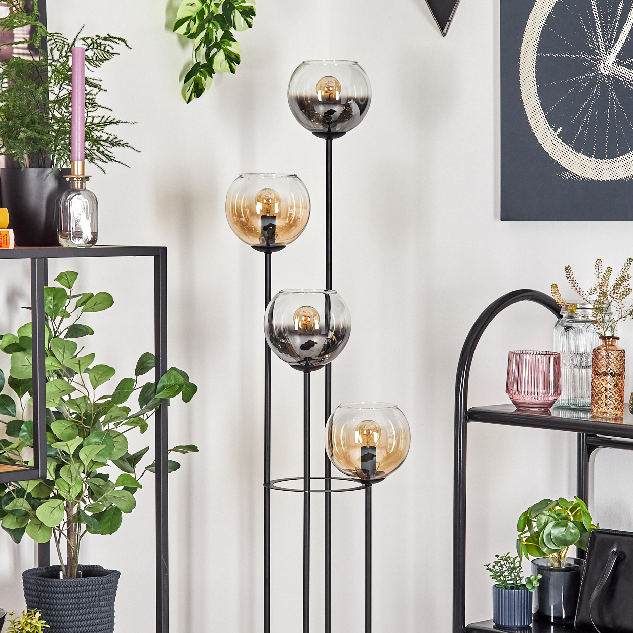 Eine moderne Stehlampe mit vier Glaskugelschirmen. Die Umgebung umfasst Pflanzen, Regale und Kunstwerke. Die Lampe hat einen schwarzen Rahmen.