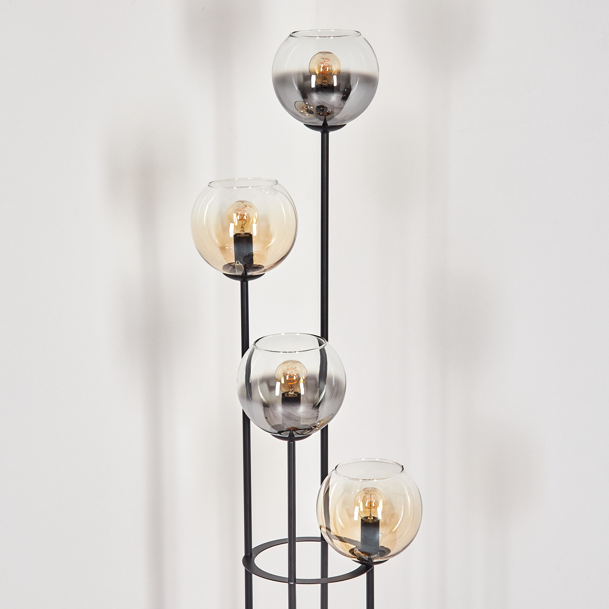 Eine moderne Stehlampe mit vier Glasschirmen, jede mit einer Glühbirne, auf einem schwarzen Metallrahmen, vor einer weißen Wand.