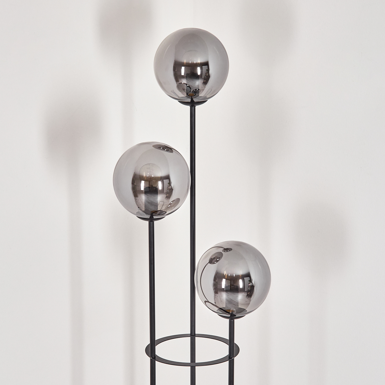 Eine moderne Lampe mit drei kugelförmigen, verspiegelten Glasschirmen auf schwarzen Metallstielen vor einer weißen Wand.