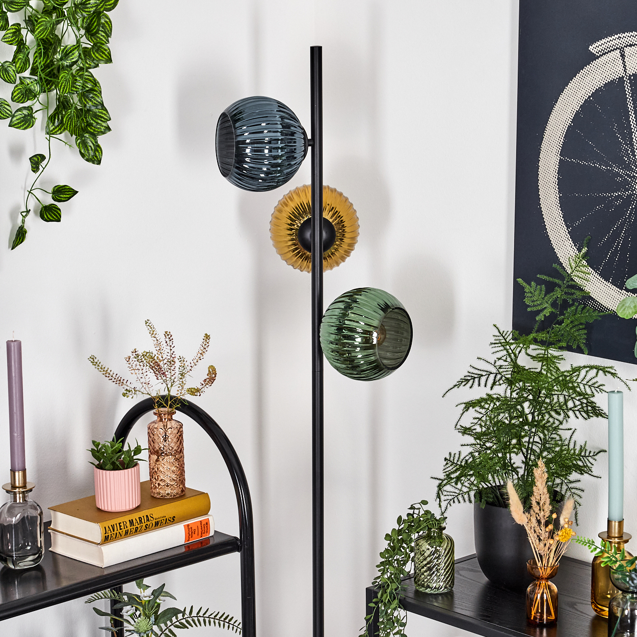 Eine moderne Stehlampe mit drei Glasschirmen. Der Raum hat Pflanzen, Regale mit Büchern und Vasen, ein Poster und Kerzen.