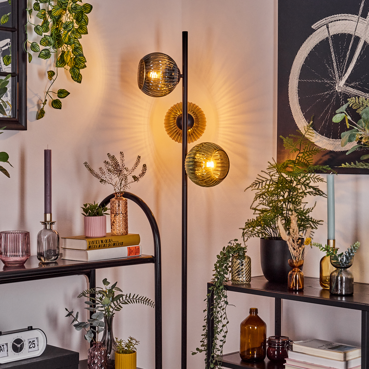 Raumecke mit Stehlampe, Pflanzen, Büchern und Kerzen auf Regalen, sowie einem Fahrradrad-Kunstwerk an der Wand.