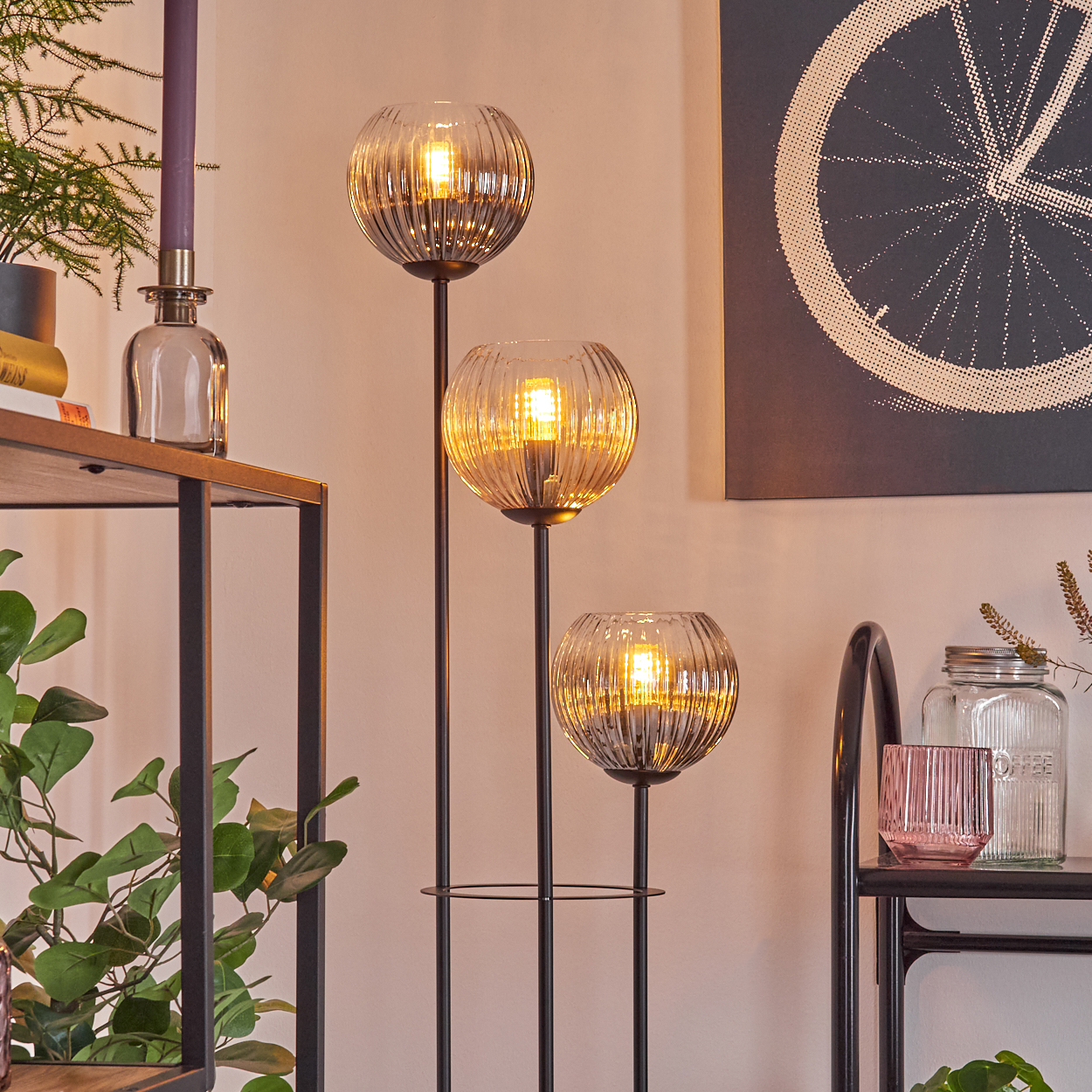 Eine dreiflammige Lampe mit Glaskugeln. Dahinter ein Regal mit Pflanzen und Kerze. An der Wand ein Fahrradradbild.