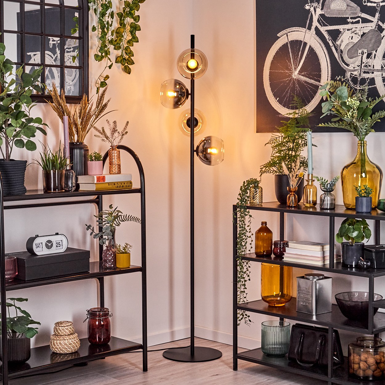 Eine Zimmerecke mit schwarzen Regalen, Pflanzen, Lampen und Dekorationsgegenständen. Ein Fahrrad-Druck befindet sich an der Wand.