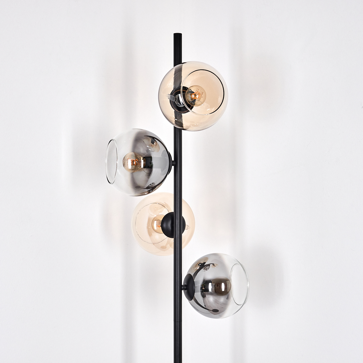 Eine moderne Stehlampe mit vier kugelförmigen Glasschirmen in verschiedenen Bernstein- und Grautönen, befestigt an einem schwarzen Metallpfosten, vor einer weißen Wand.