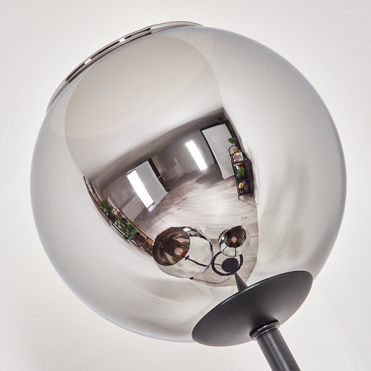 Eine reflektierende, kugelförmige Lampe. Die Kugel reflektiert einen Raum mit Fenstern, Möbeln und Pflanzen. Der Sockel ist schwarz.
