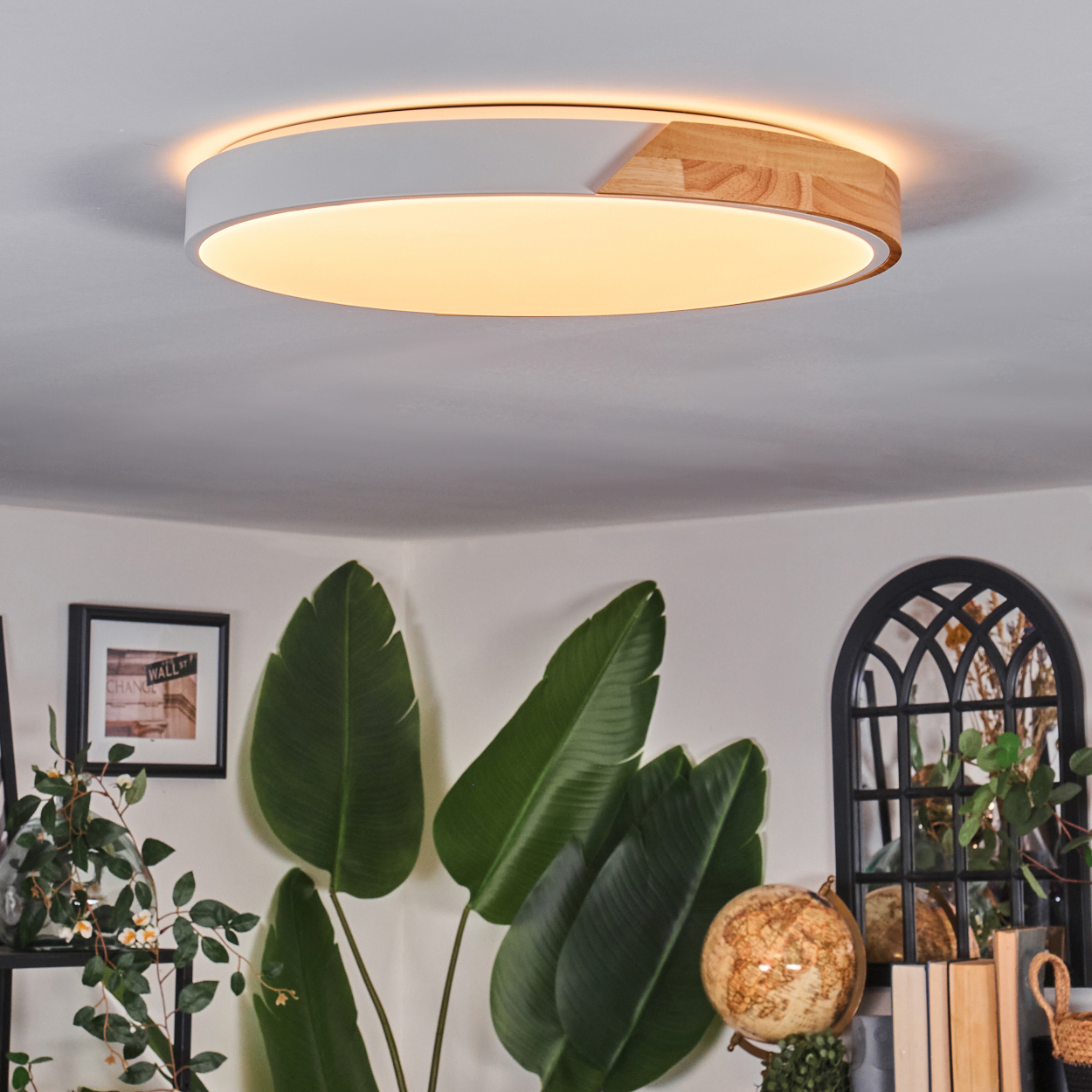 Eine moderne Deckenleuchte mit weißem und Holzfinish. Grüne Pflanzen und ein Globus sind im Vordergrund. Ein gerahmtes Bild an der Wand.