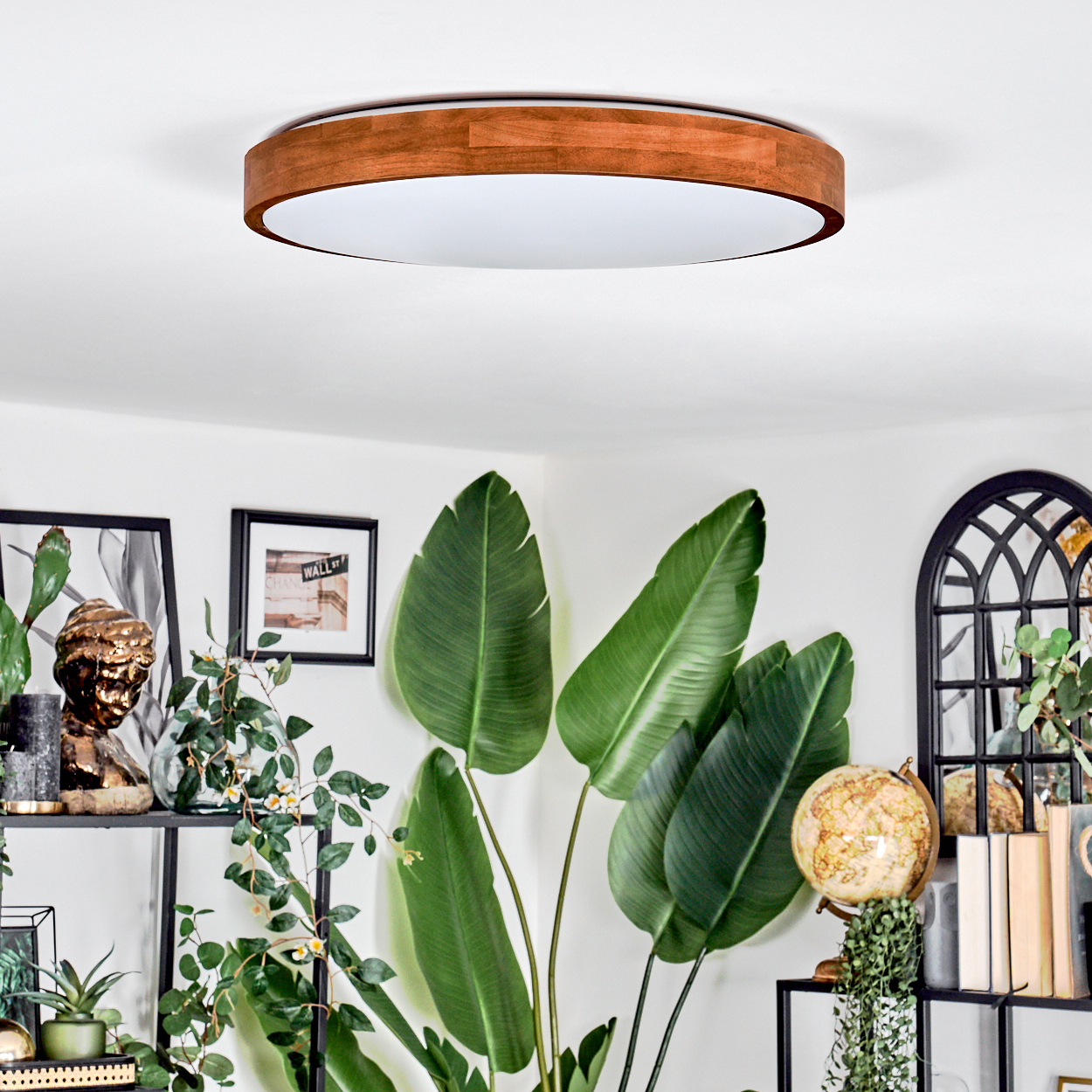 Ein modernes Wohnzimmer mit einer Holzdeckenlampe und großen grünen Pflanzen. Rahmen und ein Globus stehen auf Regalen. Weiße Wände.