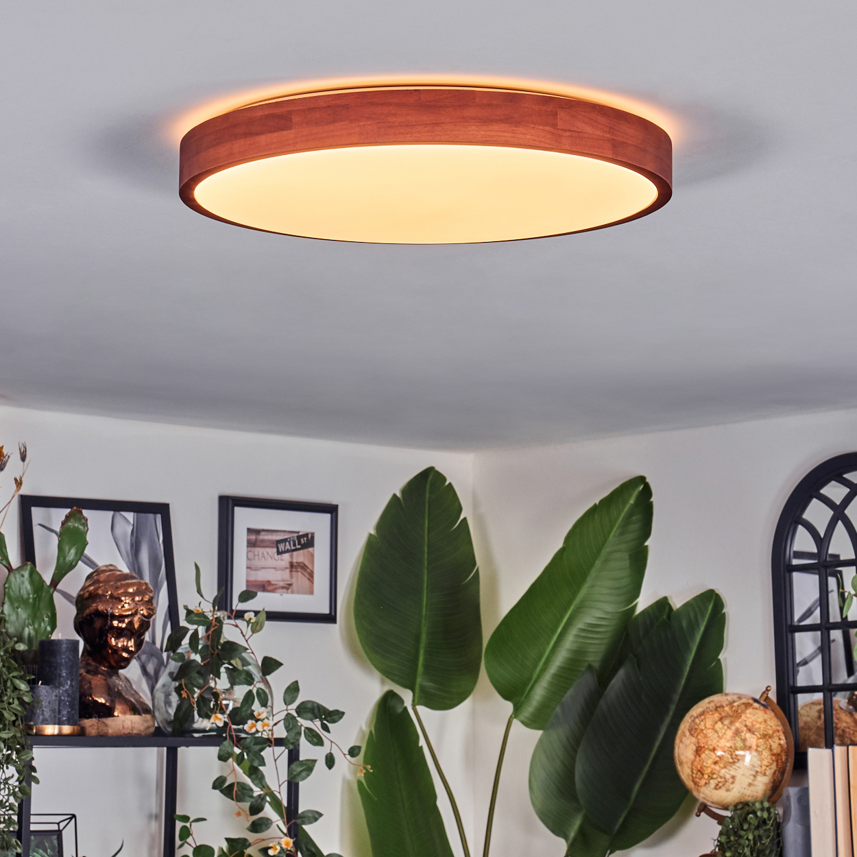Eine runde Deckenlampe aus Holz beleuchtet einen Raum mit Pflanzen, Kunstwerken, einem Globus und einer Büste. Das Licht ist warm.