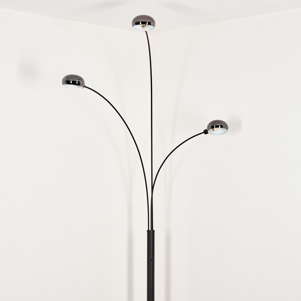 Eine moderne schwarze Stehlampe mit drei gebogenen Armen, die jeweils ein Chromlicht halten. Sie steht vor einer weißen Ecke.