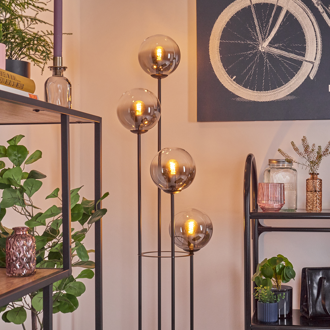 Eine Stehlampe mit vier Glaskugelleuchten steht in der Nähe von Regalen mit Pflanzen und Dekoration. Ein Fahrradkunstdruck hängt an der Wand.