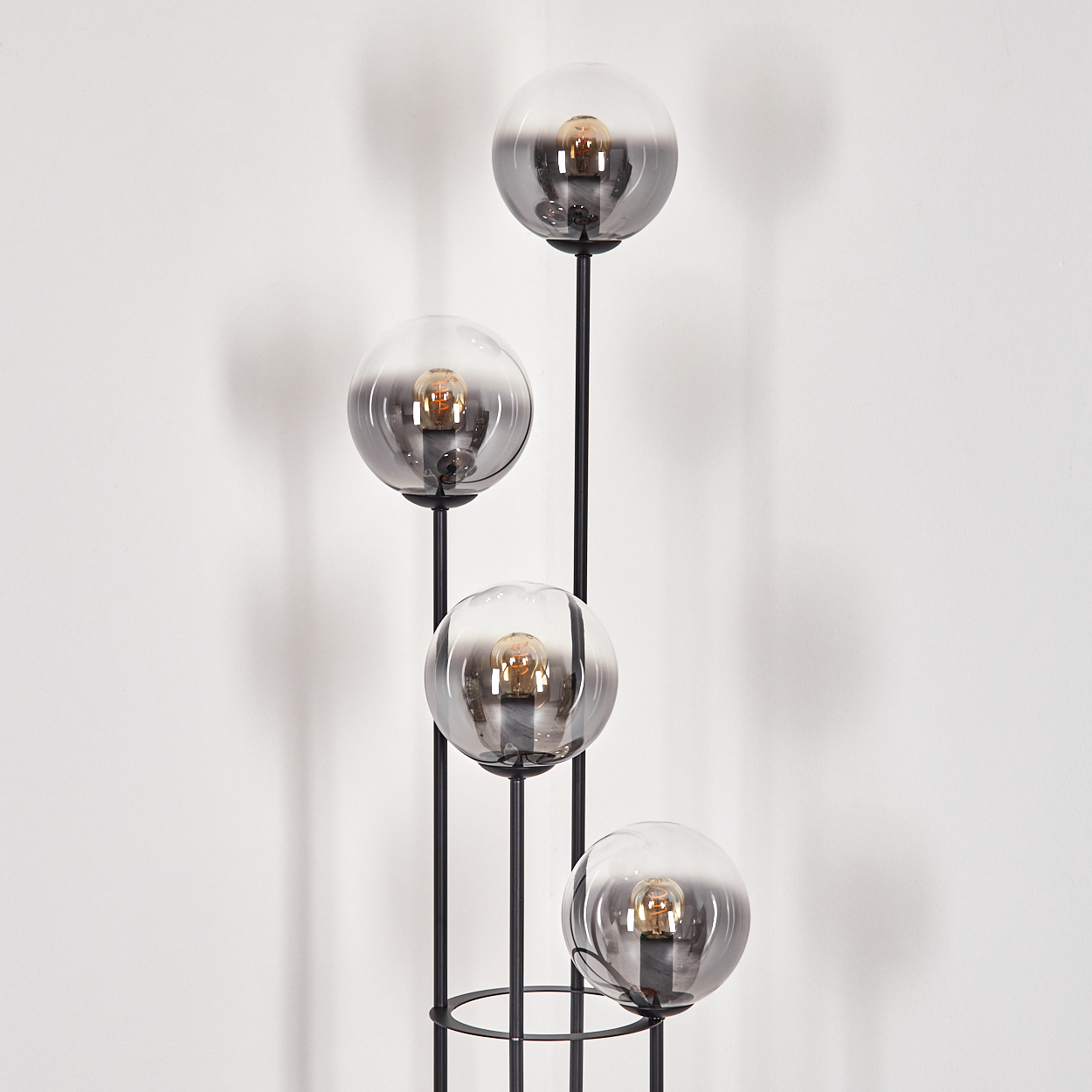 Eine moderne schwarze Stehlampe mit vier kugelförmigen, rauchfarbenen Glasschirmen, jede mit einer warmen Glühbirne, vor einer weißen Wand.