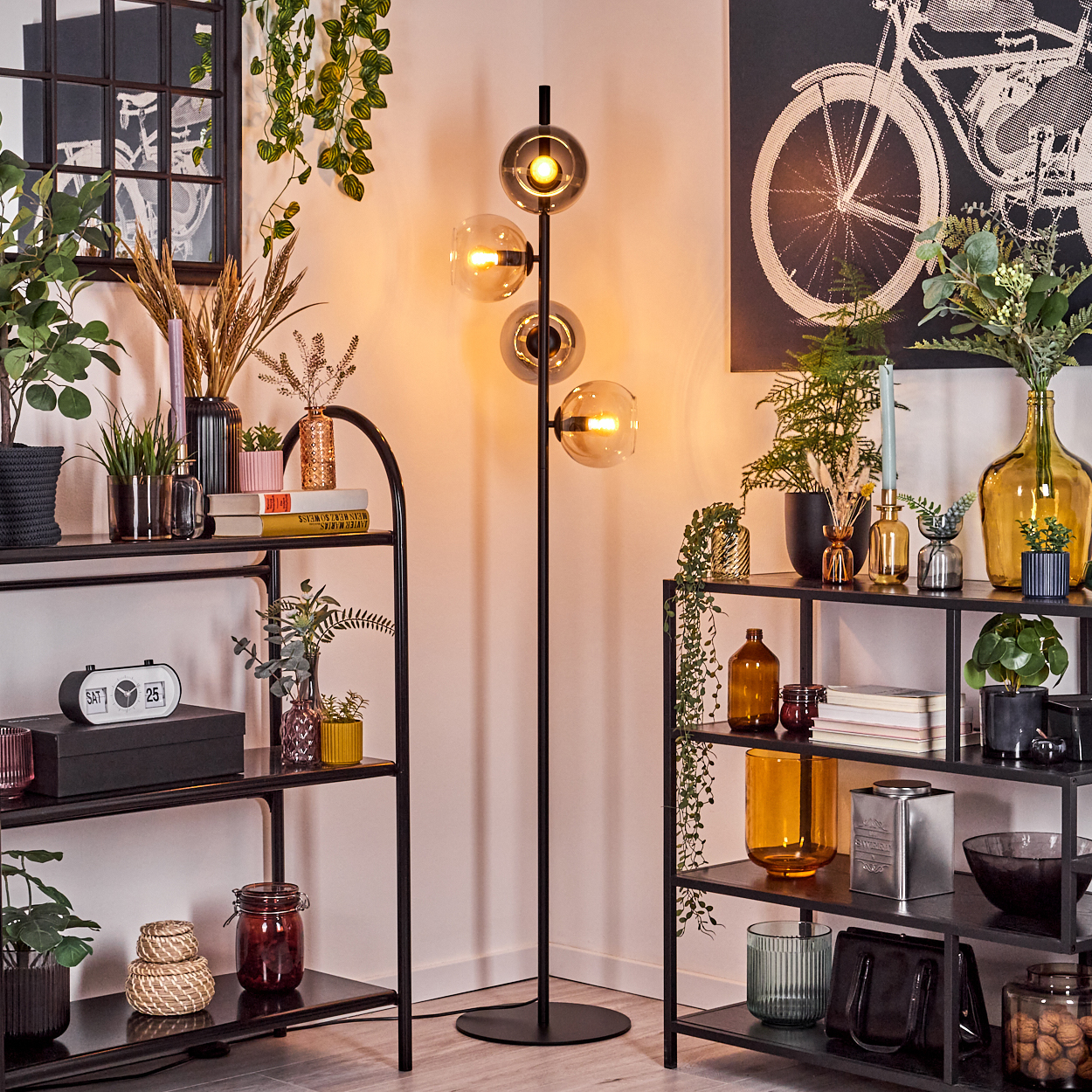 Ecke eines Wohnzimmers mit schwarzen Regalen, einer Stehlampe, Pflanzen und Dekorationsartikeln. Ein Fahrrad-Kunstdruck hängt an der Wand.