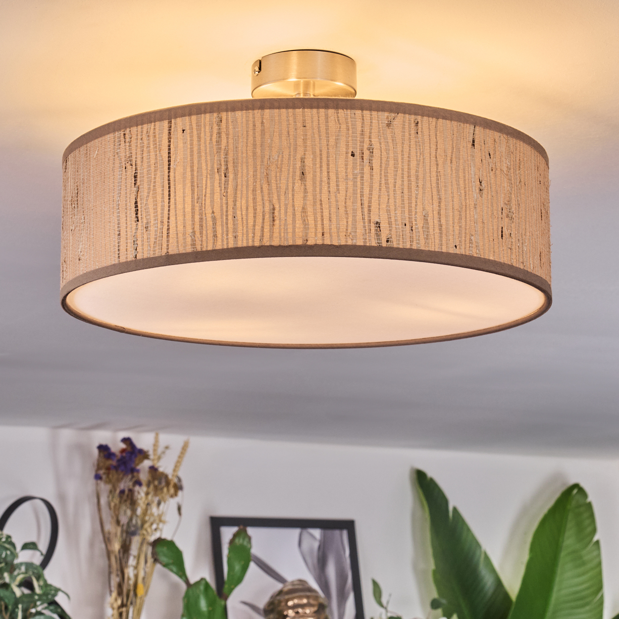 Ein beigefarbener Lampenschirm hängt von der Decke. Pflanzen und Kunstwerke sind unter der Leuchte zu sehen. Das Licht ist an.