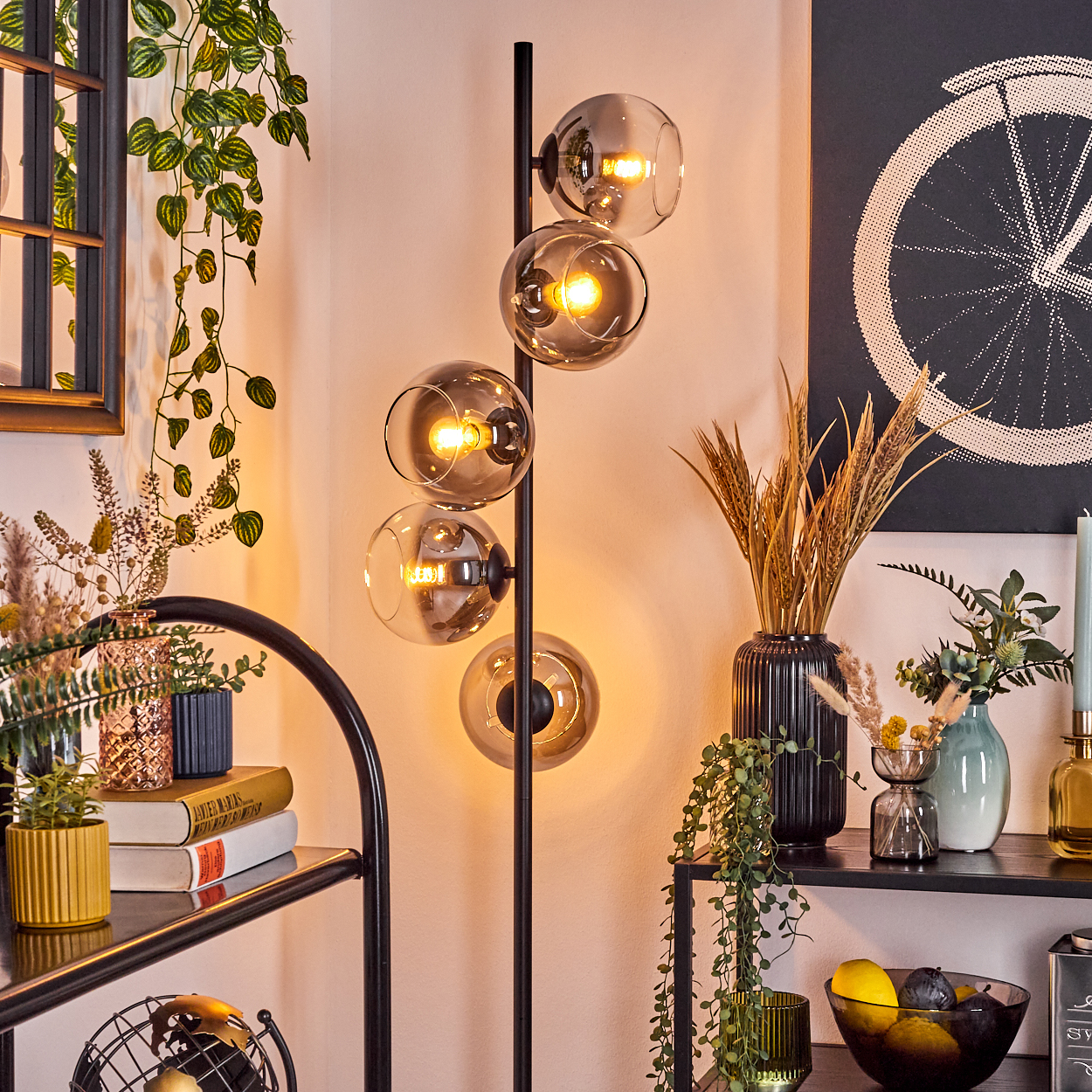Eine moderne Stehlampe mit vier Glaskugelschirmen beleuchtet einen Raum mit verschiedenen Pflanzen, einem schwarzen Regal und Dekorationsartikeln.