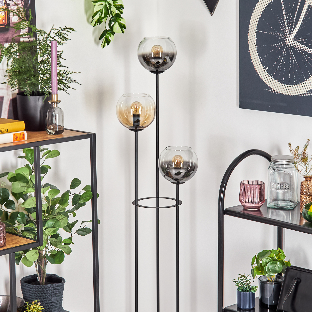 Eine Stehlampe mit drei Glaskugeln. Auf beiden Seiten Regale mit Pflanzen und Dekoration. Ein Fahrradrad-Kunstwerk an der Wand.