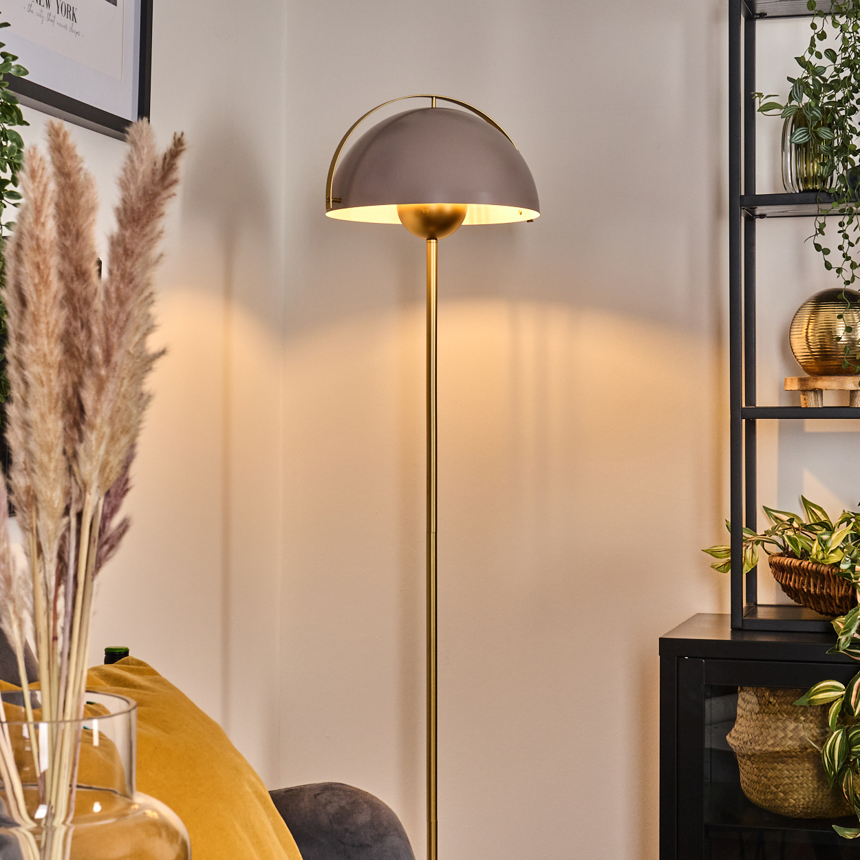 Eine moderne Stehlampe mit grauem Lampenschirm. Pflanzen stehen auf einem Regal und in einer Vase. Ein gelbes Kissen ist sichtbar.