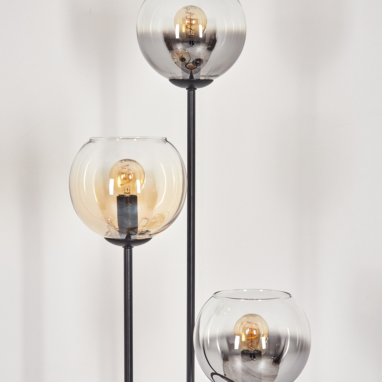 Eine Stehlampe mit drei Glaskugeln. Jede Kugel hat eine Glühbirne. Die Stiele sind schwarz vor einer weißen Wand.