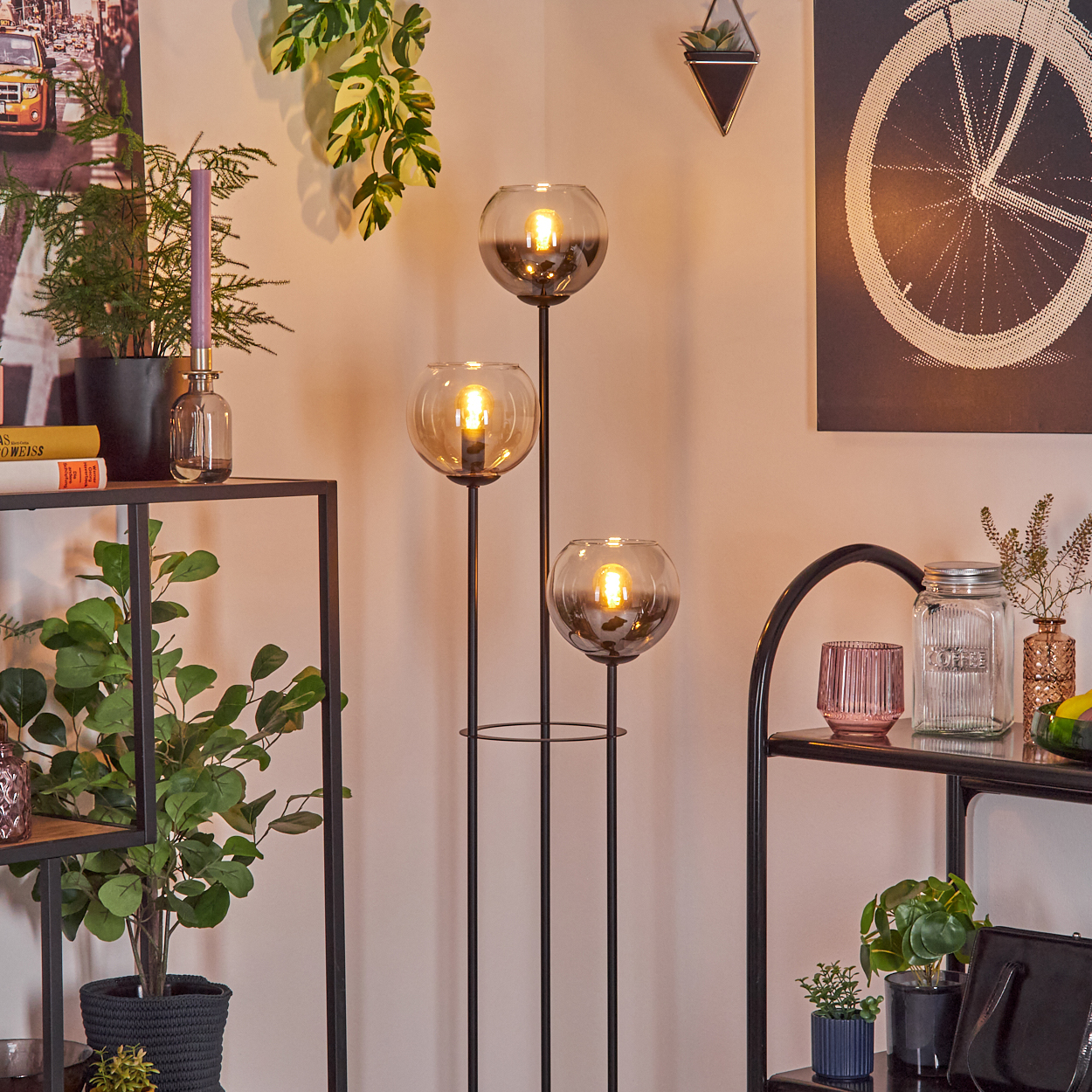 Moderne Wohnecke mit Stehlampe und drei Glaskugeln. Regale zeigen Pflanzen und Dekor, Fahrradkunst an der Wand.