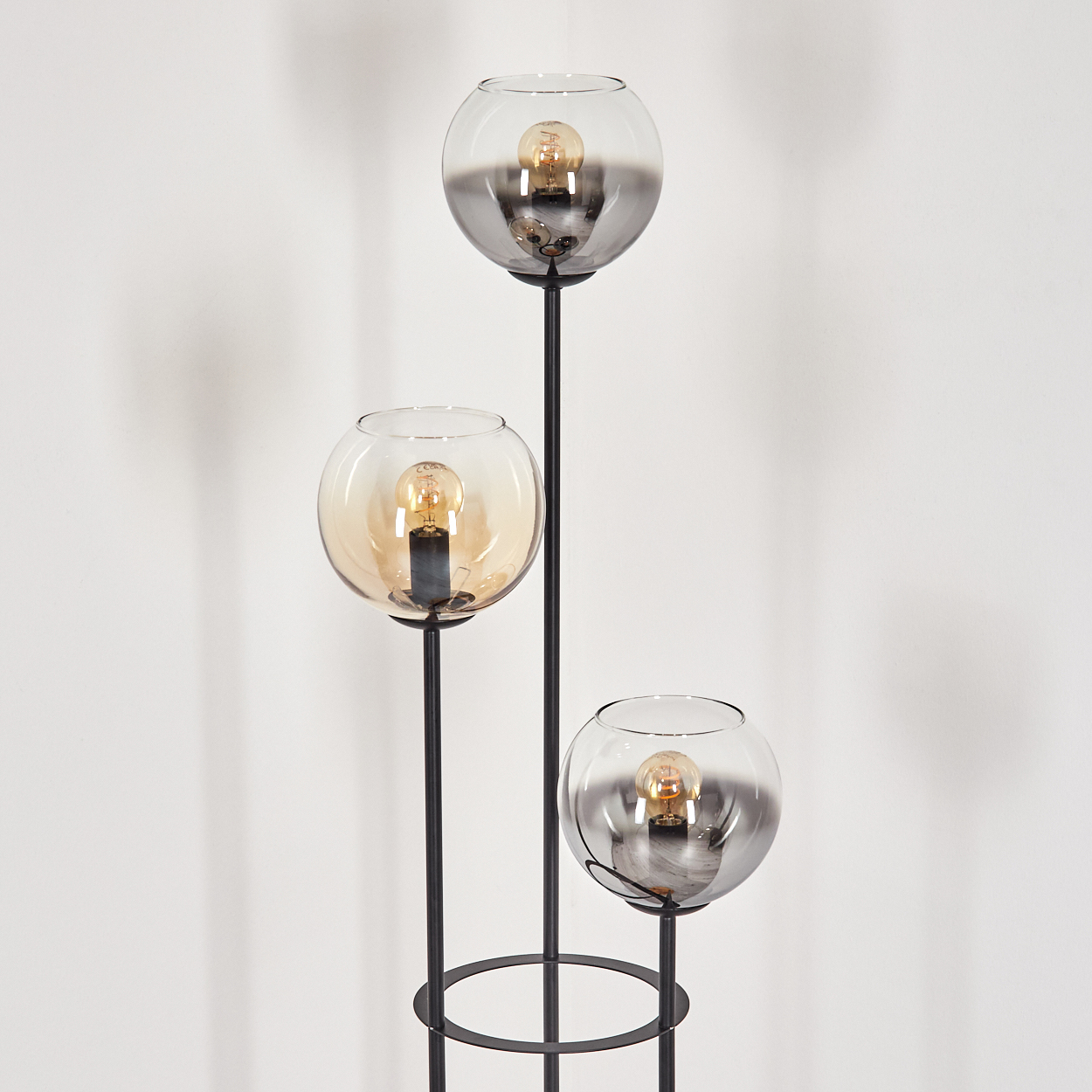 Eine moderne Stehlampe mit drei kugelförmigen Glasschirmen, die jeweils eine Glühbirne enthalten. Der Rahmen ist schwarz, vor einer weißen Wand.