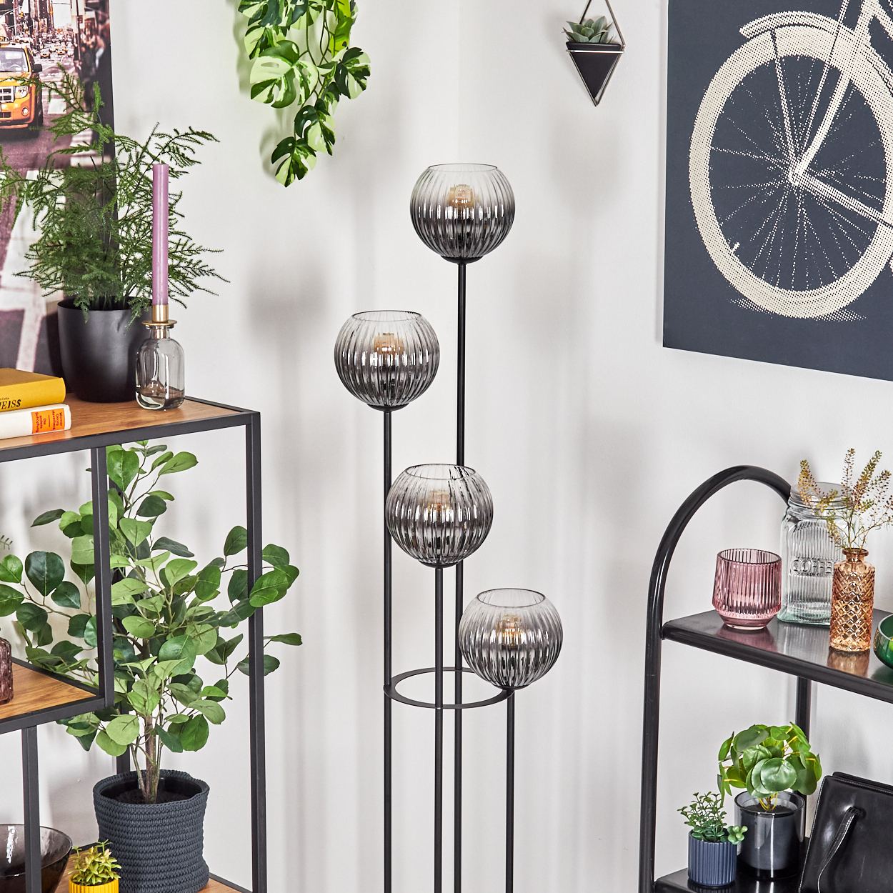 Eine dekorative Lampe mit vier Glaskugeln, Pflanzen auf Regalen und ein Fahrradrad-Kunstwerk an einer weißen Wand.